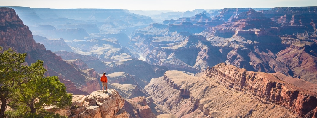 A male hiker is standing on a steep cliff taking in the amazing view over famous Grand Canyon on a beautiful sunny day with blue sky in summer, Grand Canyon National Park, Arizona, USA