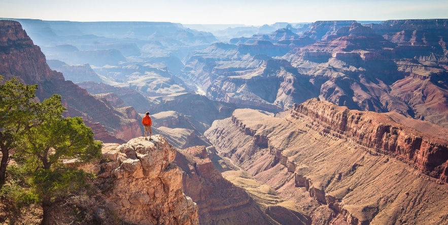 A male hiker is standing on a steep cliff taking in the amazing view over famous Grand Canyon on a beautiful sunny day with blue sky in summer, Grand Canyon National Park, Arizona, USA