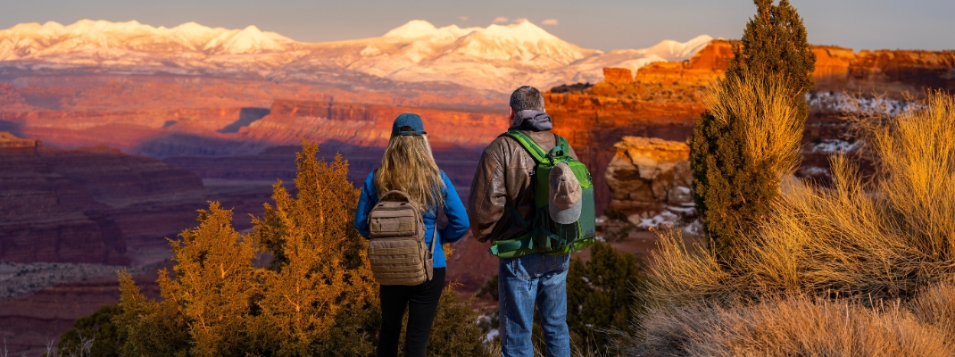 Couple enjoying beautiful sunset in winter in the mountains in Utah. Friends relaxing on hiking trip. Canyonlands National Park at sunset against snow covered mountains. Utah, USA. 