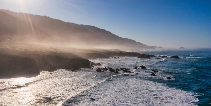 Aerial view of Big Sur coastline along Highway 1 in California, with ocean waves, rocky shoreline, and morning mist illuminated