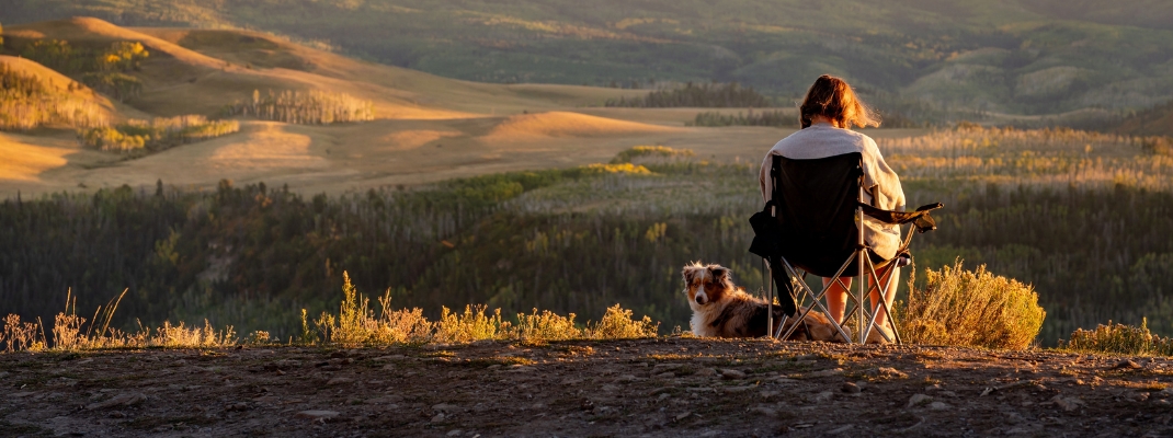 Female traveler sitting on a camping chair with her dog watching the view of San Juan mountains during sunset